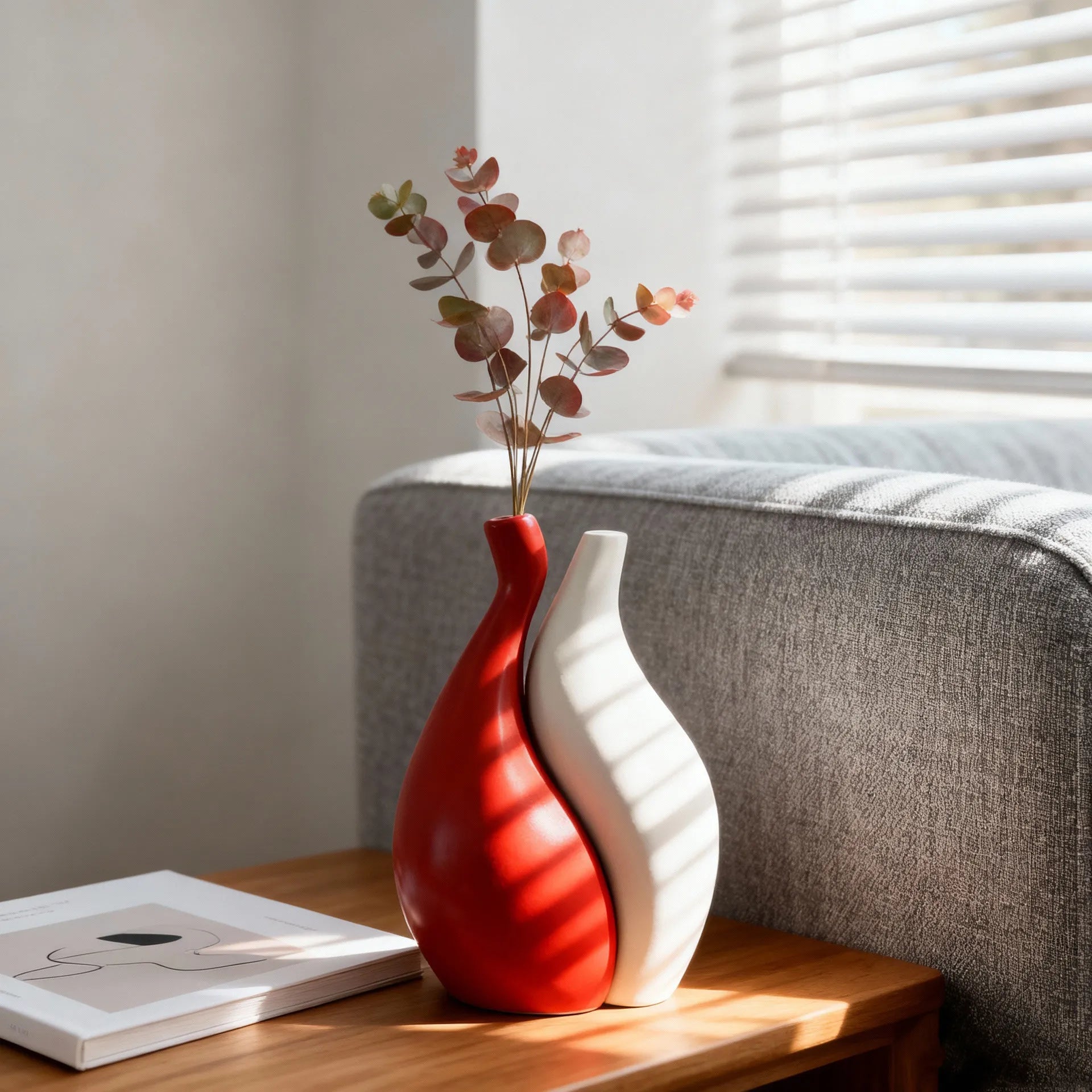 Two modern ceramic vases, one glossy red and one matte white, arranged side by side on a wooden table next to a gray sofa with eucalyptus branches in the red vase.